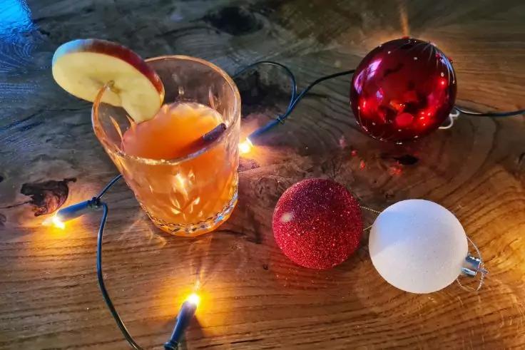 Auf Holztisch: Glas mit orangefarbener Fl&uuml;ssigkeit und Apfelscheibe am Rand, Lichterkette mit warmen Lichtern und drei Kugeln (rot gl&auml;nzend, rot glitzernd, wei&szlig;)