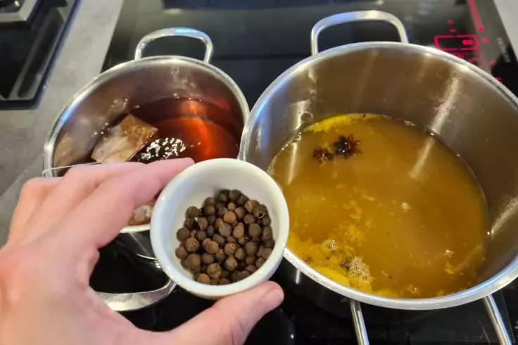 Hand h&auml;lt eine kleine wei&szlig;e Sch&uuml;ssel mit ganzen Pfefferk&ouml;rnern &uuml;ber zwei T&ouml;pfen auf einem Herd; rechter Topf enth&auml;lt helle braune Fl&uuml;ssigkeit mit einem Sternanis, linker Topf enth&auml;lt dunkle braune Fl&uuml;ssigkeit mit einem Beutel