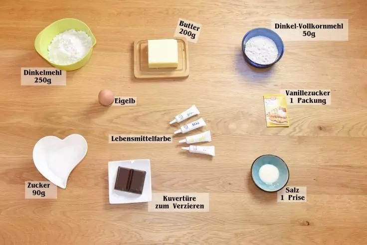 Auf einem Holztisch angeordnet: grüne Schale mit weißem Pulver, Butterblock auf Holzbrett, kleine blaue Schale mit hellem Mehl, Päckchen mit Aufschrift 'Vanillezucker', herzförmige weiße Schale mit Zucker, kleine Schale mit Eigelb, mehrere Tuben Lebe