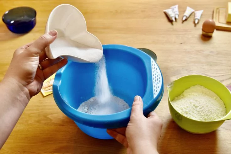 Zwei Hände schütten weißes Pulver aus einer herzförmigen weißen Schale in eine blaue Rührschüssel auf einem Holztisch; daneben eine grüne Schüssel mit weißem Pulver, ein Ei, kleine Tuben und eine dunkle Schale