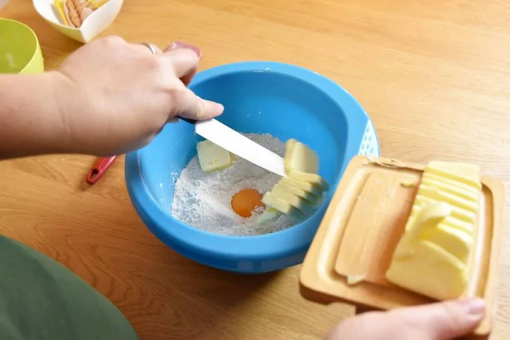Zwei Hände schaben mit einem Messer Butter von einer Butterdose in eine blaue Schüssel mit Mehl und einem rohen Ei auf einem Holztisch