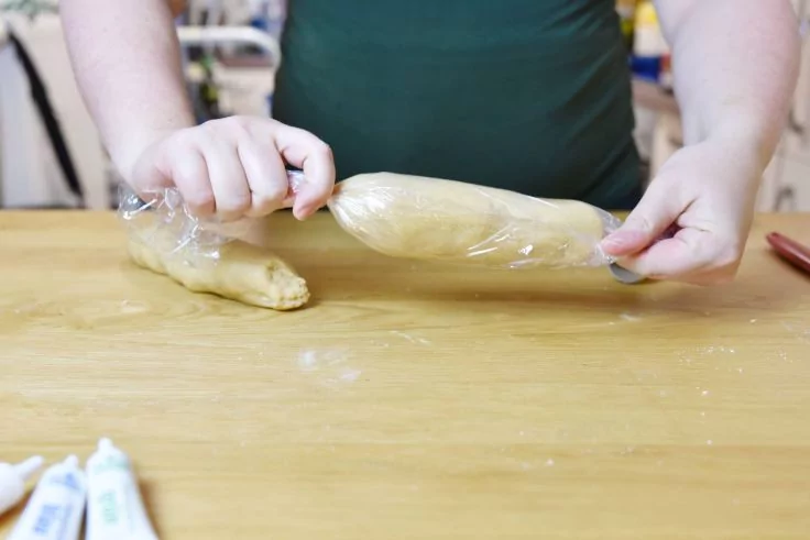 Hände halten eine in Frischhaltefolie gewickelte zylindrische Teigrolle auf einem bemehlten Holzbrett; weitere Teigrolle, drei Tuben im Vordergrund, unscharfe Küchenutensilien im Hintergrund
