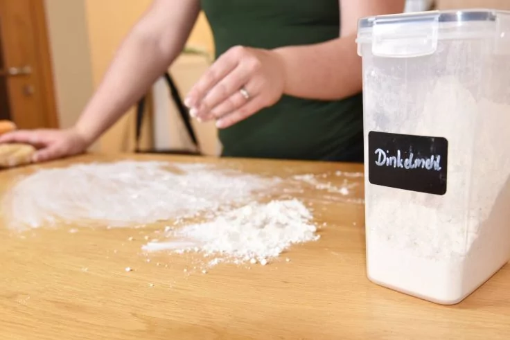 Hände einer Person bearbeiten Teig auf einem Holztisch, Mehl auf der Arbeitsfläche, rechts ein Kunststoffbehälter mit Aufkleber 'Dinkelmehl'