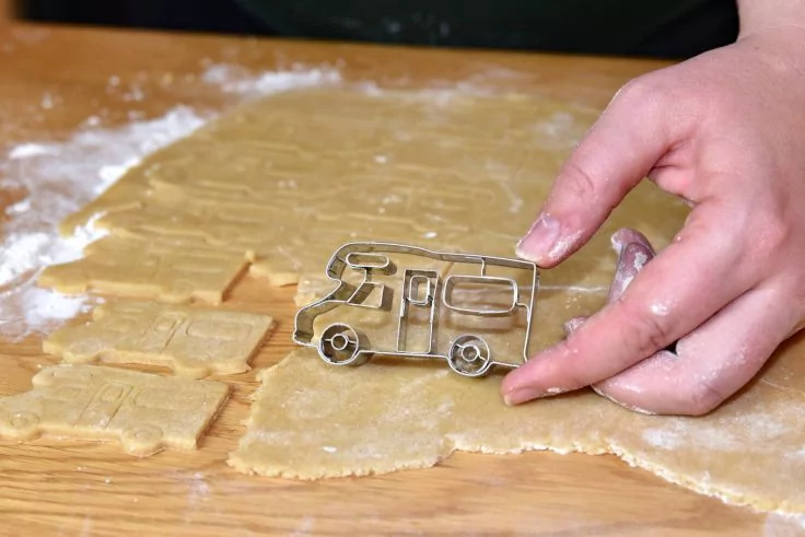 Hand drückt Metall-Ausstecher in Form eines Wohnmobils in ausgerollten Teig auf einem Holzbrett, Mehl und ausgestochene Teigstücke sichtbar