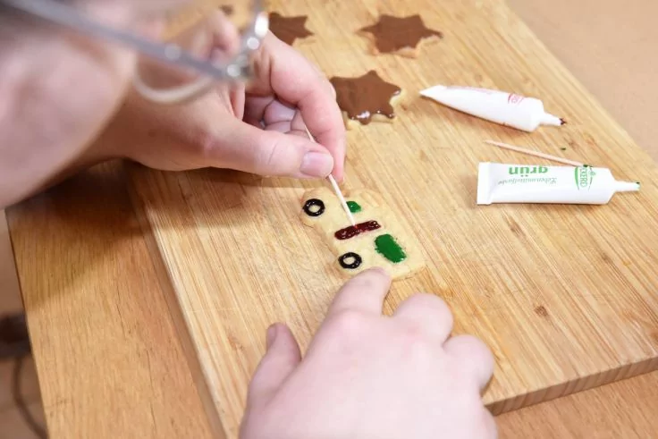 Zwei Hände halten einen Zahnstocher und bemalen kleine Formen (grüne Rechtecke, schwarze Ringe) auf einem Holzbrett; zwei kleine Tuben und braune sternförmige Stücke liegen daneben