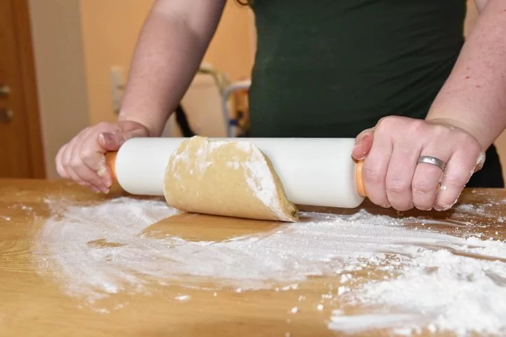 Hände und Oberkörper einer Person, die Teig mit einem Nudelholz auf einer bemehlten Holzfläche ausrollt