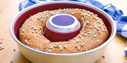 Ringf&ouml;rmiges Brot mit Sonnenblumenkernen in runder Backform auf Holzuntergrund, blau-wei&szlig; kariertes Tuch im Hintergrund