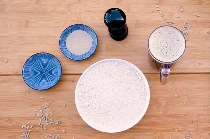 Gro&szlig;e wei&szlig;e Sch&uuml;ssel mit wei&szlig;em Pulver, zwei kleine blaue Schalen (eine mit Pulver), kleines schwarzes Gef&auml;&szlig;, Glasbecher mit hellem schaumigem Inhalt, verstreute K&ouml;rner auf hellem Holzuntergrund