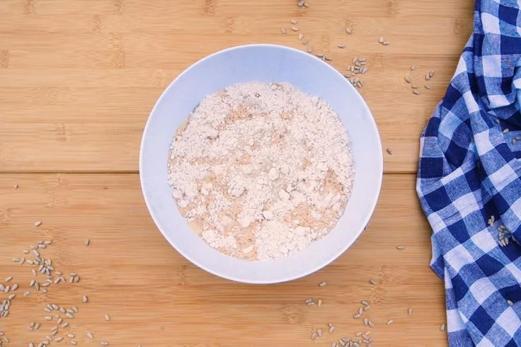 Draufsicht auf eine wei&szlig;e Sch&uuml;ssel mit trockenem, feinem hellbraunem Pulver auf hellem Holzbrett; einzelne hellbraune Samen um die Sch&uuml;ssel; rechts ein gefaltetes blau-wei&szlig; kariertes Tuch