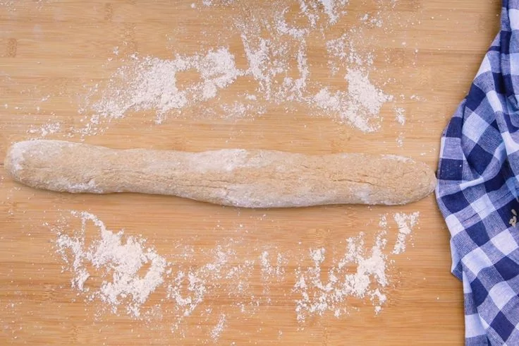 L&auml;nglich geformter Teig auf bemehltem Holzbrett, verstreutes Mehl und blau-wei&szlig; kariertes Tuch rechts