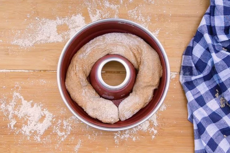 Ringf&ouml;rmiger Teig in einer Gugelhupf-Backform auf einem Holzbrett mit verstreutem Mehl, rechts ein blau-wei&szlig; kariertes Tuch