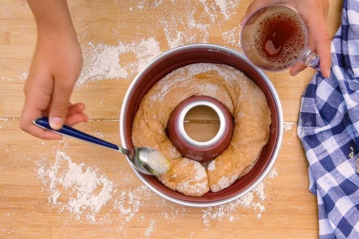 Vogelperspektive: zwei H&auml;nde &uuml;ber einer runden Kranz-Backform mit mehreren runden Teigst&uuml;cken, eine Hand h&auml;lt einen L&ouml;ffel, die andere ein kleines Glas mit brauner Fl&uuml;ssigkeit; Mehl auf Holzfl&auml;che, blau-wei&szlig; kariertes Tuch rechts