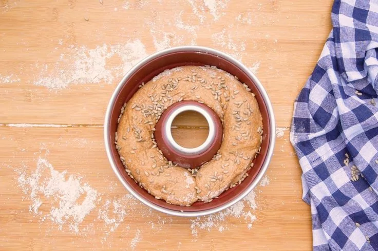Ringf&ouml;rmiges Brot mit Samen in einer Backform auf Holzbrett, verstreutes Mehl und blau-wei&szlig; kariertes Tuch rechts