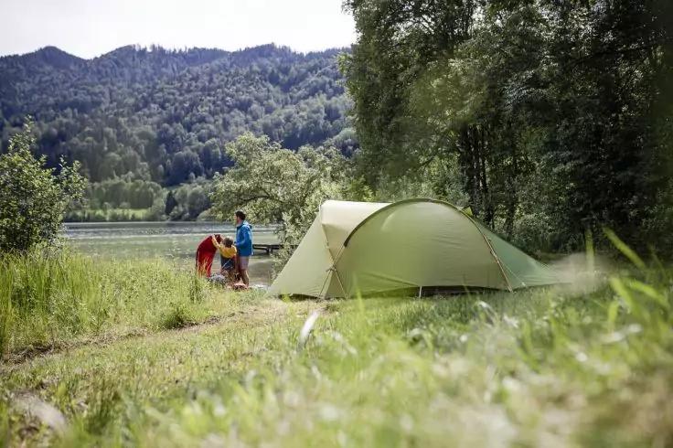 Gr&uuml;nes Zelt am Seeufer, zwei Personen neben dem Zelt, Gras im Vordergrund, B&auml;ume und bewaldete H&uuml;gel im Hintergrund