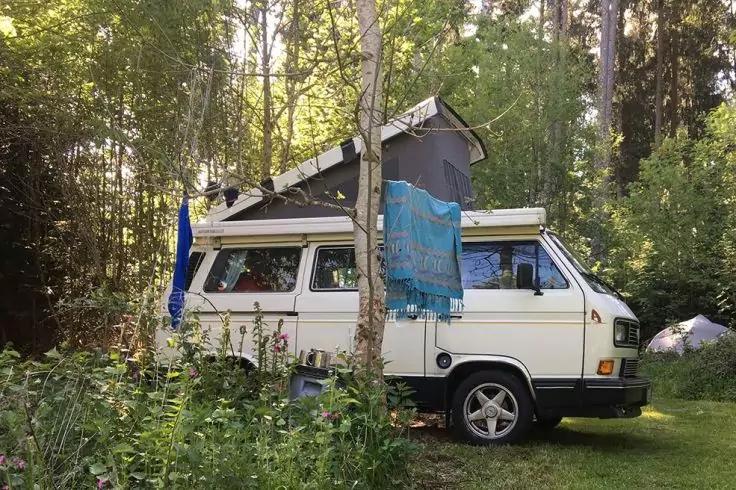 Wei&szlig;er Camper mit aufgestelltem Aufstelldach, blaues Tuch h&auml;ngt an der Seite, geparkt auf Gras, umgeben von B&auml;umen