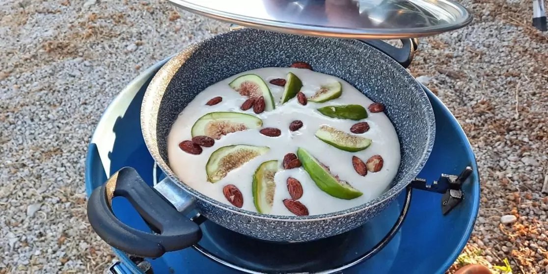 Grauer, marmorierter Topf mit schwarzen Griffen auf blauem Campingkocher, Deckel halb offen. Im Topf wei&szlig;liche Fl&uuml;ssigkeit mit gr&uuml;nen Fruchtspalten mit rosa Innen und verstreuten braunen ovalen St&uuml;cken. Kiesboden im Hintergrund
