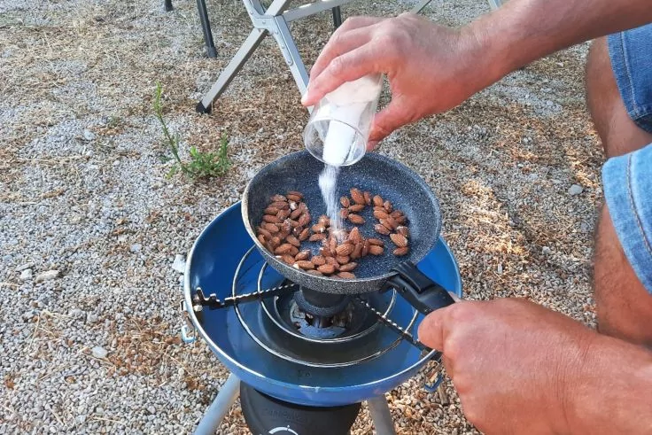 Zwei Hände schütten weißes Granulat aus einem kleinen Glas auf braune Nüsse in einer grauen Pfanne über einem blauen Campingkocher auf kiesigem Boden; im Hintergrund ein Klappstuhl
