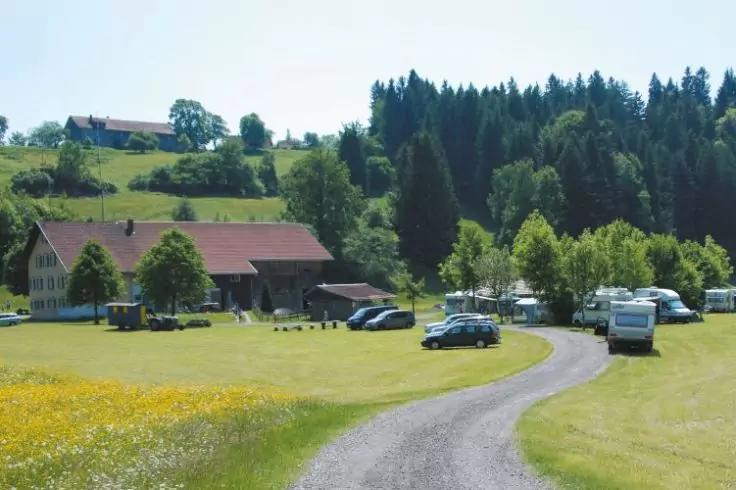 Ein geschotterter Weg f&uuml;hrt durch eine gr&uuml;ne Wiese mit gelben Blumen zu geparkten Autos und Wohnmobilen; links ein gro&szlig;es Geb&auml;ude mit rotem Dach, im Hintergrund Wald und H&uuml;gel