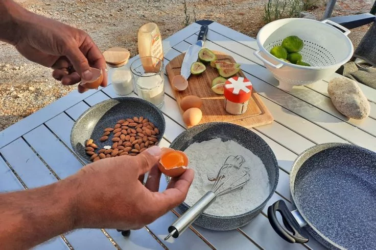 Hände schlagen ein Ei über einer Schüssel mit Mehl auf einem Tisch auf; auf dem Tisch Pfannen mit Mandeln, eine leere Pfanne, Eier, ein Schneebesen, halbierte Feigen auf einem Holzbrett, ein Sieb mit Limetten und kleine Behälter