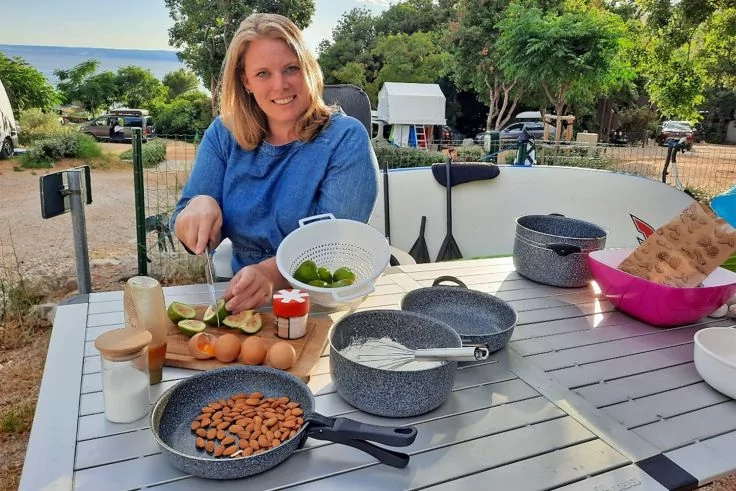 Person sitzt an einem Tisch im Freien und schneidet Gemüse; auf dem Tisch Pfannen mit Mandeln, Eier, Gläser, ein Sieb mit Limetten und Kochgeschirr; im Hintergrund Bäume, Zelte und das Meer