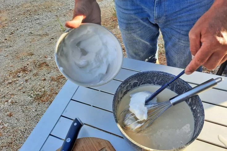Hände über einem weißen Tisch: eine hält eine Metallschüssel mit weißer Creme, die andere rührt in einer beschichteten Pfanne mit einem Schneebesen; Messer auf Holzbrett, Person trägt blaue Jeans, kiesiger Boden im Hintergrund