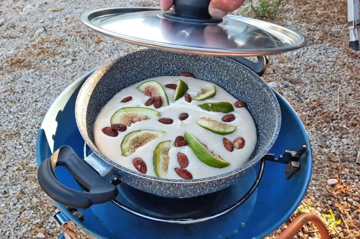 Eine Hand hebt den Deckel von einer grauen, gesprenkelten Pfanne auf einem blauen Campingkocher; in der Pfanne eine weiße Flüssigkeit mit halbierten grünen Feigen und ganzen braunen Mandeln; Kiesboden im Hintergrund