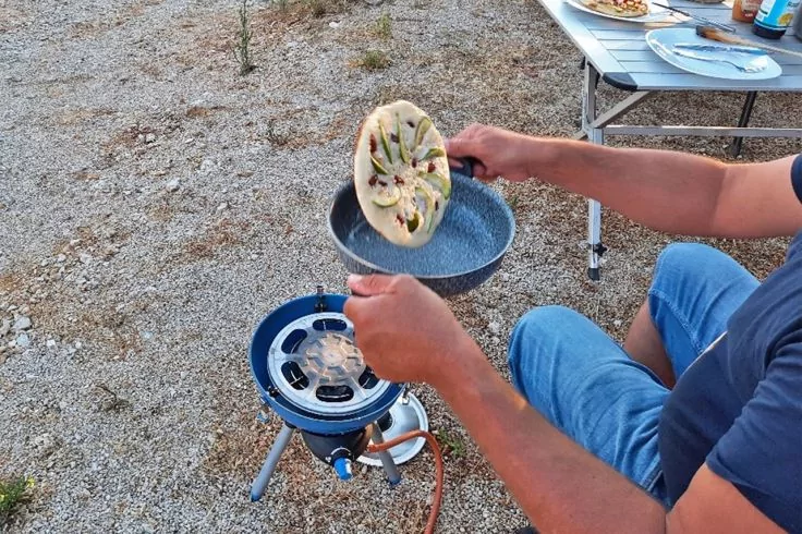Eine Person sitzt im Freien und hält eine Pfanne mit einem rund belegten Fladenbrot über einem blauen Campingkocher; im Hintergrund ein Klapptisch mit Tellern und Kiesboden
