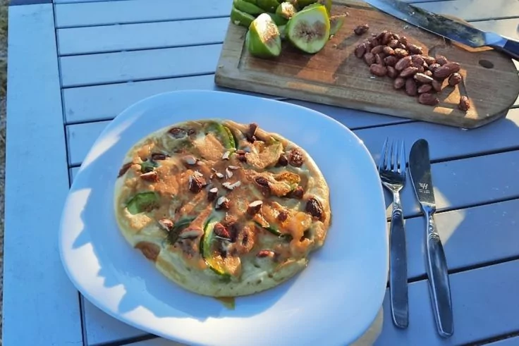 Weißer Teller mit rundem, goldbraunem Gericht belegt mit grünen Scheiben und Nüssen; rechts Besteck; im Hintergrund Holzbrett mit geschnittenem grünem Obst und Nüssen auf blauem Latten-Tisch