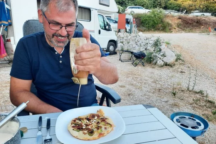 Mann sitzt an einem Tisch im Freien und drückt eine Saucenflasche über eine kleine Pizza auf einem Teller; Wohnmobil und Campingstühle im Hintergrund