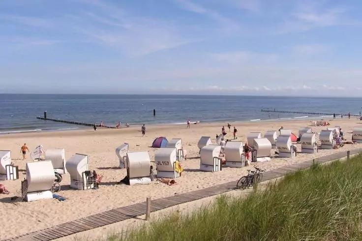 Sandstrand mit einer Reihe wei&szlig;er Strandk&ouml;rbe am Ufer, mehrere Personen auf dem Sand, Holzsteg und Meer unter blauem Himmel