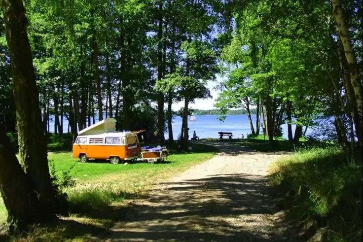 Orange Wohnmobil auf einer Wiese unter B&auml;umen, kleiner Campingtisch mit zwei St&uuml;hlen daneben, Schotterweg zum See, Picknickbank am Ufer, See im Hintergrund