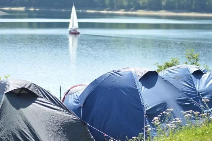 Mehrere blaue Zelte am Ufer, im Hintergrund ein Segelboot auf dem See