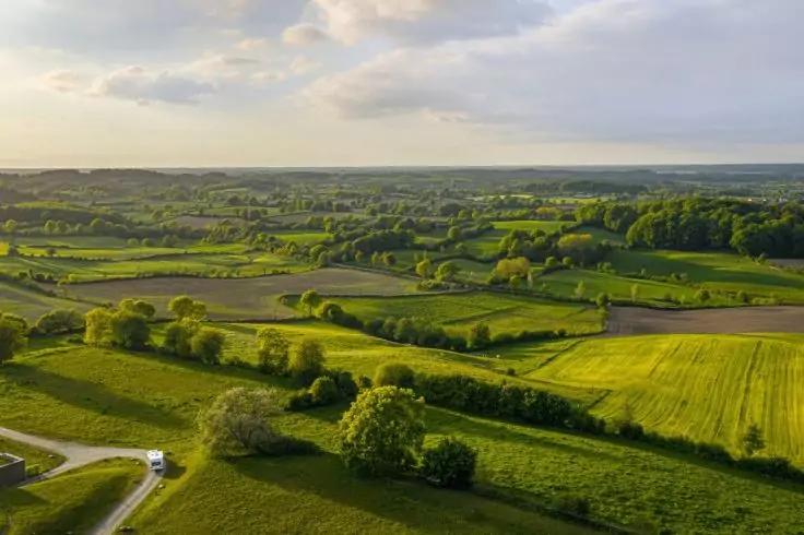 Luftaufnahme h&uuml;geliger gr&uuml;ner Felder, getrennt durch Hecken und Baumreihen, schmale Stra&szlig;e mit einem wei&szlig;en Fahrzeug, bew&ouml;lkter Himmel