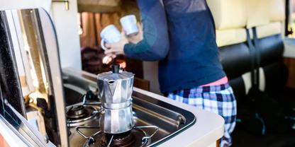 Mokkakanne auf Gasherd in Wohnmobilk&uuml;che, daneben stehende Person in Pyjamahose h&auml;lt zwei Tassen; Sitzbank im Hintergrund