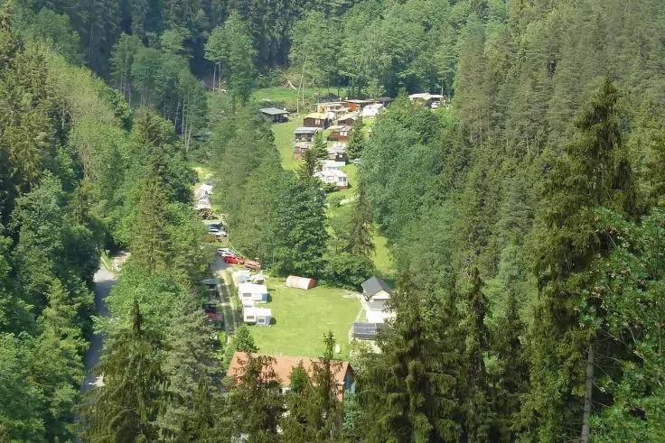 Mehrere Wohnwagen und kleine Geb&auml;ude auf einer grasbewachsenen Lichtung, umgeben von hohen Nadelb&auml;umen an einem bewaldeten Hang