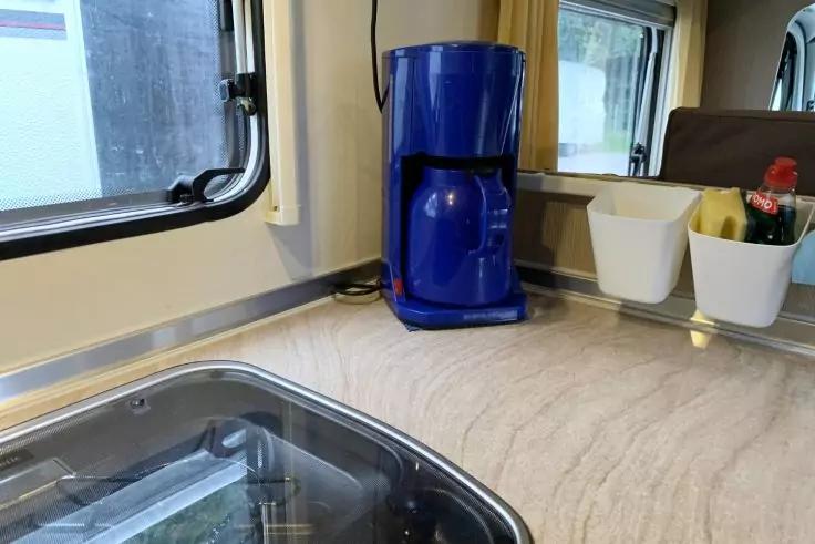 Blaue Kaffeemaschine auf heller Arbeitsplatte neben Spüle mit Glasabdeckung, links ein Fenster, rechts zwei weiße Behälter mit Schwamm und Flasche