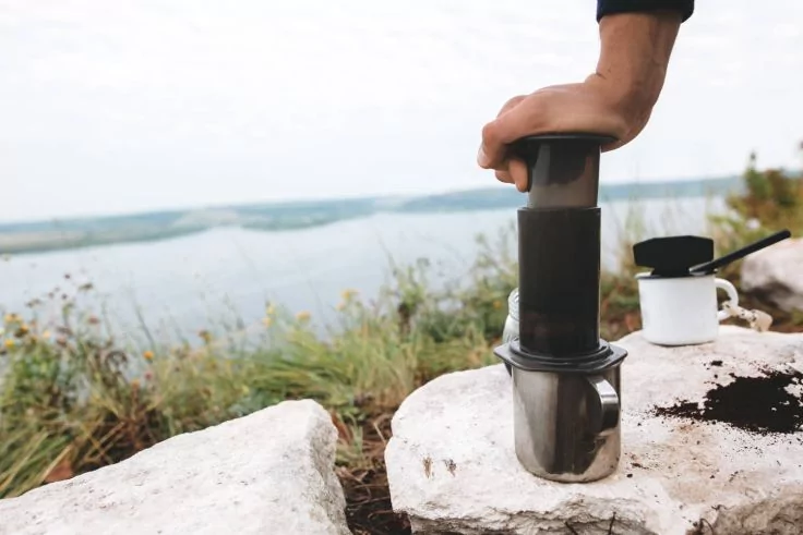 Hand drückt AeroPress auf einen Metallbecher auf einem Felsen; daneben weiße Emailletasse und verstreutes Kaffeepulver, im Hintergrund See und Ufer