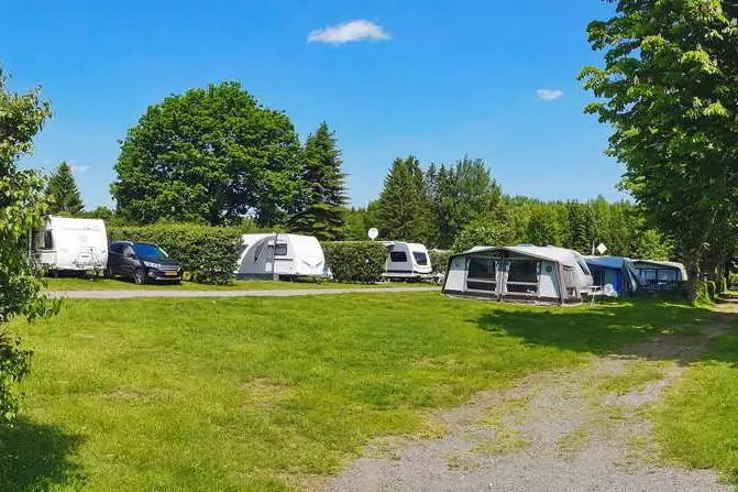 Mehrere Wohnwagen mit Vorzelten auf einem grasbewachsenen Campingplatz, B&auml;ume und blauer Himmel, schmaler Schotterweg im Vordergrund