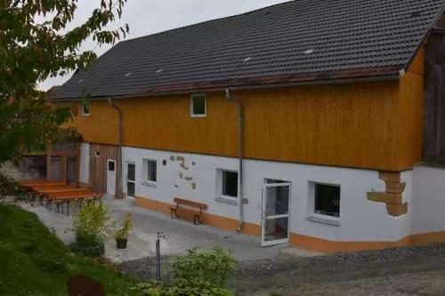 Zweist&ouml;ckiges Geb&auml;ude mit gelber Holzfassade oben, wei&szlig;er Putzfassade unten, Glast&uuml;r, Bank, Au&szlig;entische und Baum im Vordergrund