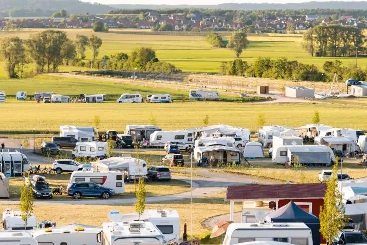 Campingplatz mit mehreren Wohnmobilen und Wohnwagen auf einem flachen Feld, im Hintergrund B&auml;ume und ein Dorf