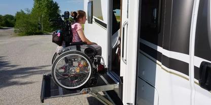 Person im Rollstuhl auf einer Plattform-Hebeb&uuml;hne vor ge&ouml;ffneter T&uuml;r eines wei&szlig;en Wohnmobils, mit Rucksack, Stra&szlig;e und B&auml;umen im Hintergrund