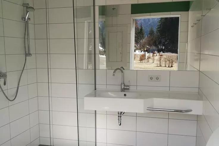 Wei&szlig; gefliestes Badezimmer mit Duschkopf links, Glasabtrennung, Waschbecken mit Armatur, Spiegel und kleinem Fenster mit Blick auf B&auml;ume und Geb&auml;ude