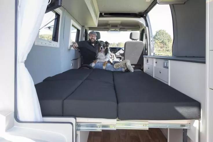 Innenraum eines Fahrzeugs mit schwarzer Schlaffl&auml;che, seitlichen Schr&auml;nken und Fenster mit Vorhang; vorn Sitze sichtbar; ein Mann sitzt auf der Schlaffl&auml;che, neben ihm liegt ein Hund