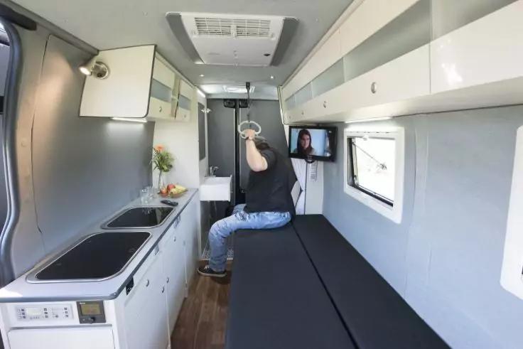 Innenraum eines Wohnmobils: linke K&uuml;chenzeile mit Sp&uuml;le und Herd, rechte lange Sitz-/Liegefl&auml;che unter H&auml;ngeschr&auml;nken, Fenster und Wandfernseher, eine Person in schwarzem Oberteil und Jeans sitzt auf der Sitzfl&auml;che und h&auml;lt eine an der Decke befestig