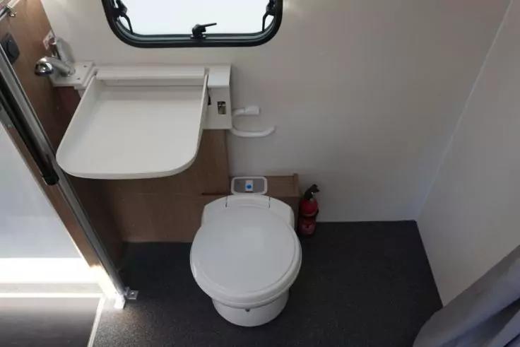 Runde wei&szlig;e Toilette auf grauem Boden, rechts ein roter Feuerl&ouml;scher, links ein klappbares wei&szlig;es Waschbecken unter einem kleinen rechteckigen Fenster mit schwarzem Rahmen