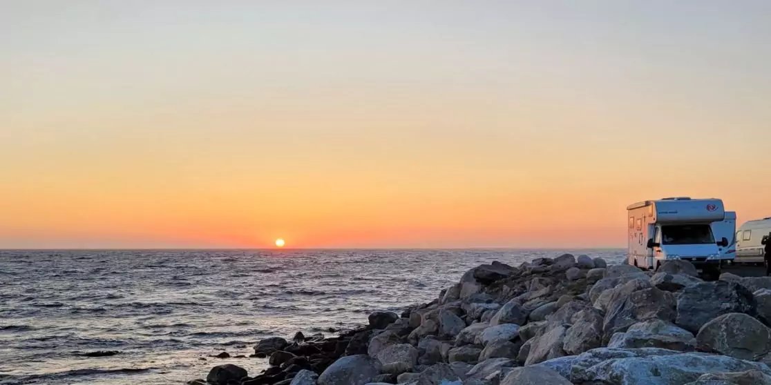 Die Sonne knapp &uuml;ber dem Horizont &uuml;ber dem Meer, Wellen, felsige K&uuml;ste im Vordergrund und ein Wohnmobil rechts auf den Felsen