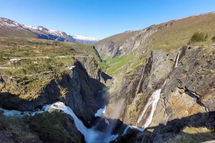 Tiefe, schmale Schlucht mit steilen Felsw&auml;nden, einem Wasserfall, der in einen Fluss st&uuml;rzt, gr&uuml;nem Tal, schneebedeckten Bergen in der Ferne und blauem Himmel