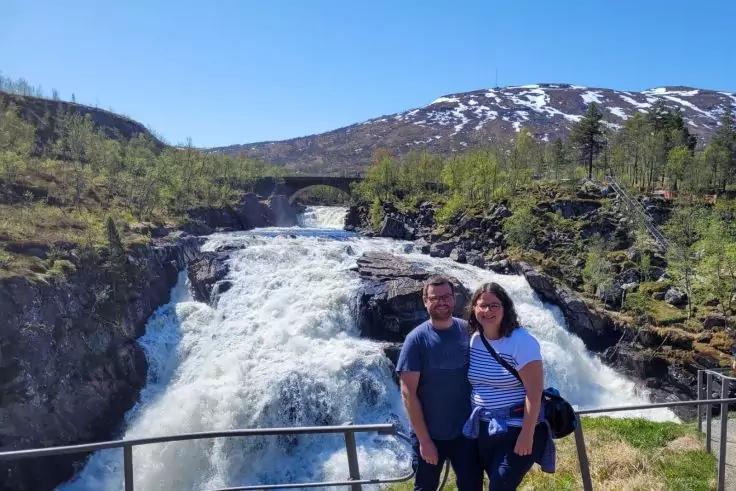 Zwei Personen stehen an einem Gel&auml;nder vor einem breiten Wasserfall; im Hintergrund eine Steinbr&uuml;cke, felsige Ufer, bewachsene H&auml;nge mit Schneeresten und blauer Himmel