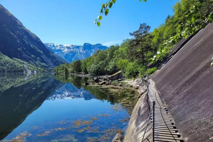 Bergsee mit spiegelnder Wasserfl&auml;che, schneebedeckte Gipfel, Baumufer und schmaler Weg mit Gel&auml;nder entlang einer Felswand
