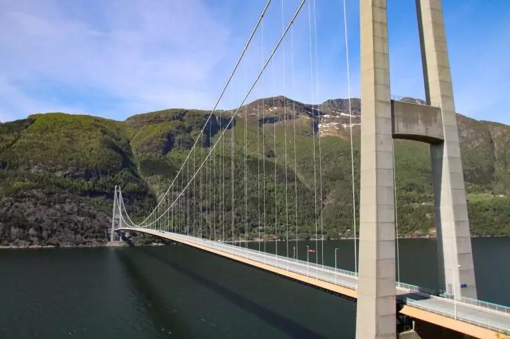 Betonbr&uuml;cke mit hohen T&uuml;rmen und gespannten Kabeln &uuml;ber einem Gew&auml;sser, im Hintergrund gr&uuml;ne Berge und blauer Himmel